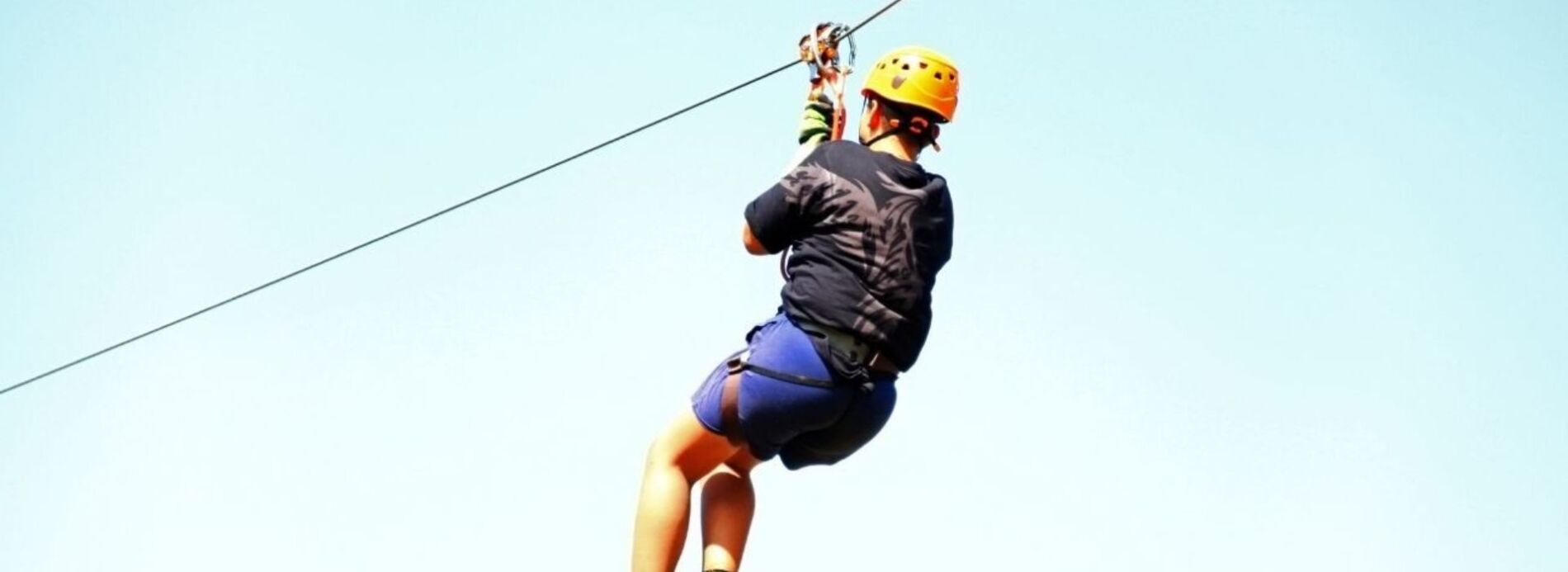 A vacationer wearing a helmet and safety harness gliding down an elevated zipline cable under a clear blue sky.
