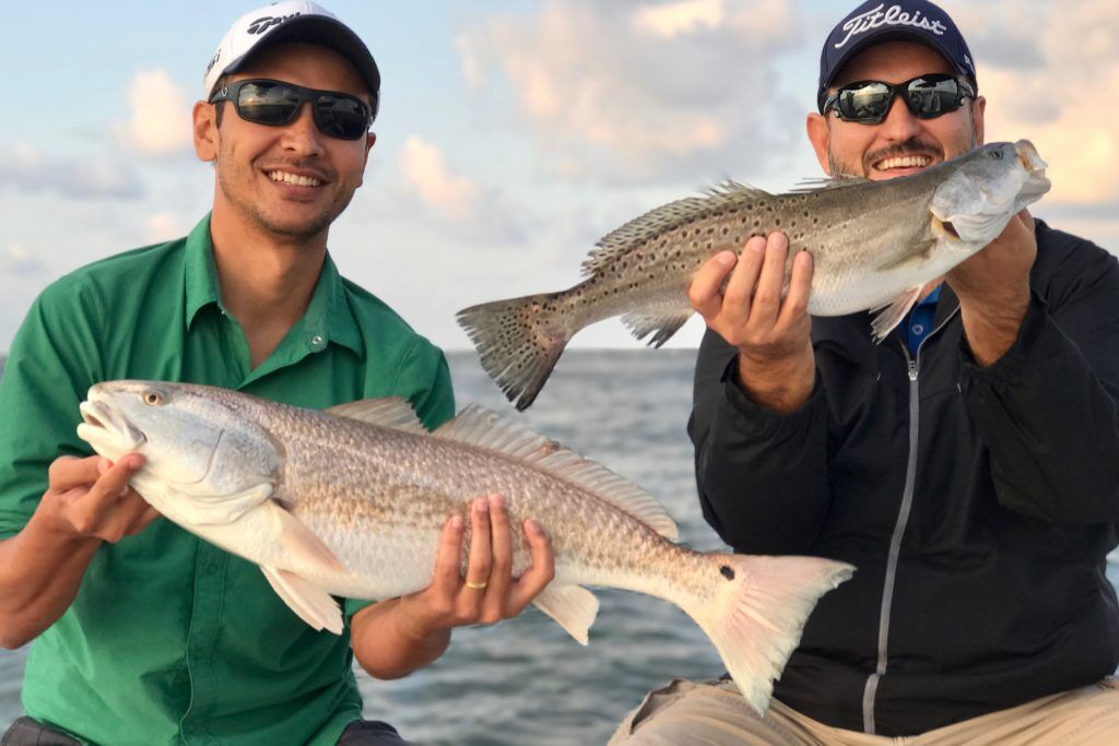 Two fishermen smiling proudly on a boat while holding up large freshly caught red drum fish, highlighting top-tier local sportfishing.