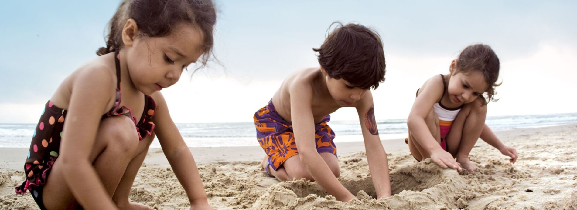 A young boy wearing a swimsuit focused on digging and playing in the soft sand during a family beach day.