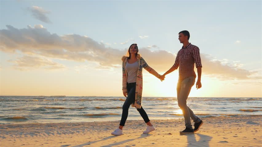 A couple holding hands and walking along the shoreline as the sun dips below the horizon, casting a warm golden glow.