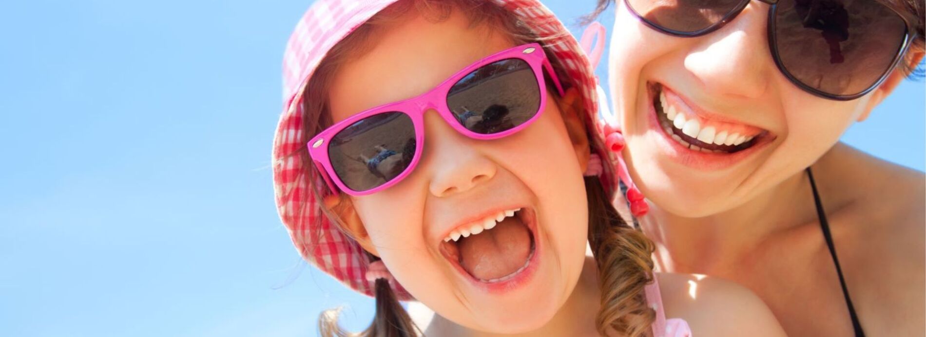 A close-up portrait of a young girl wearing bright pink sunglasses smiling excitedly during a sunny family holiday.v