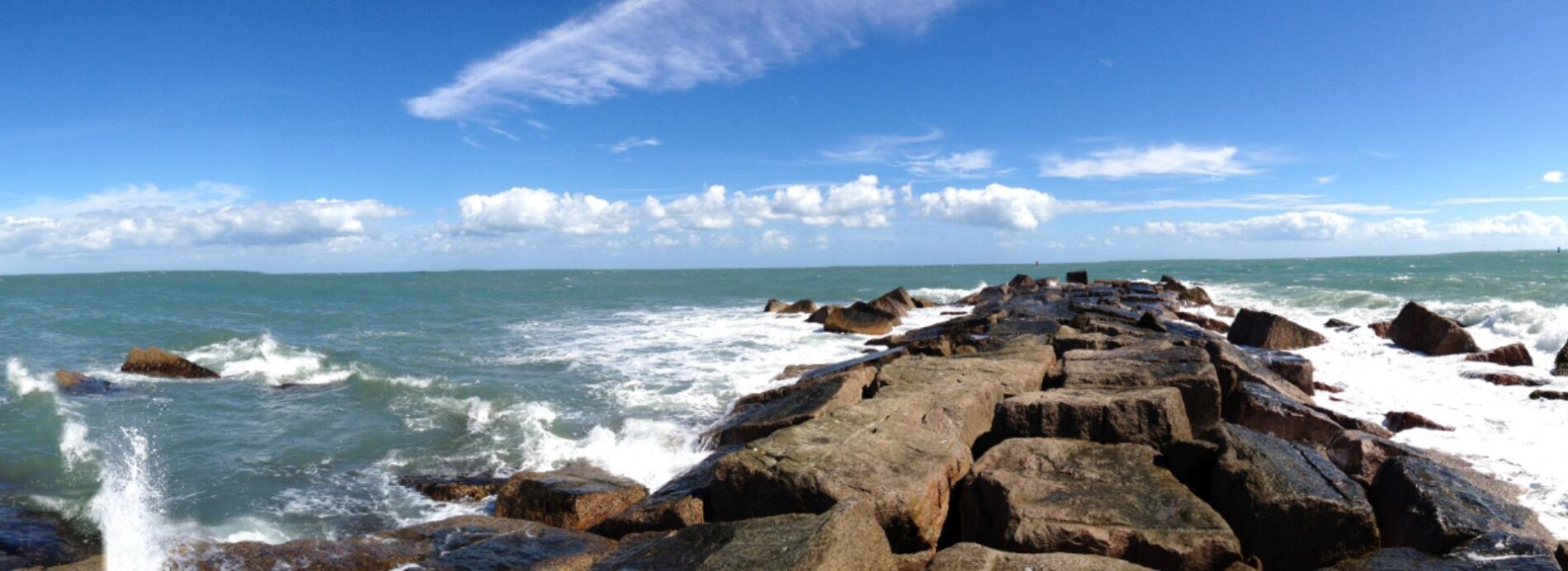 Powerful ocean waves breaking into white surf against large dark stone jetty boulders under a scattered cloudy sky.