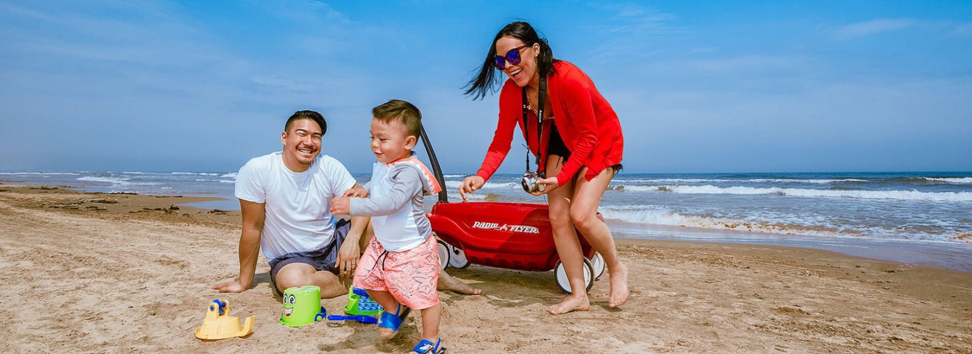 A mother pulling her young child in a classic red plastic wagon across the soft sand near the water's edge.