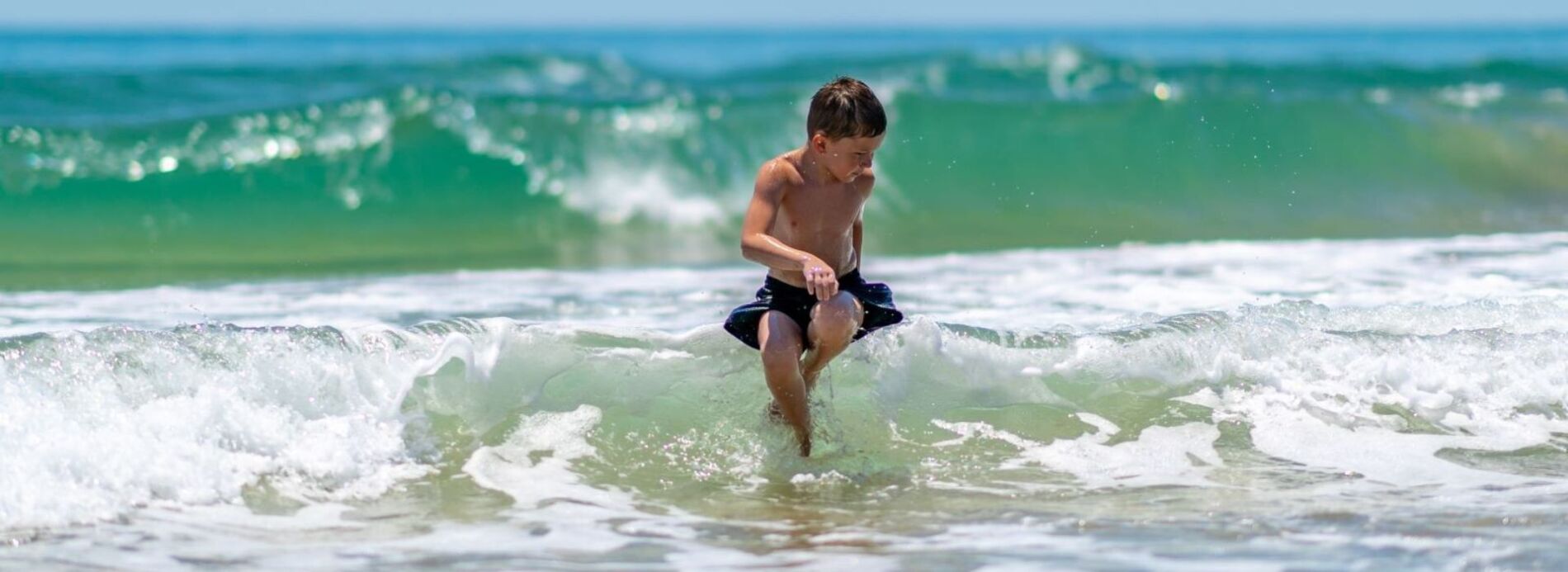 A young boy laughing as he runs through splashing ocean surf on a wide, sunny beach.