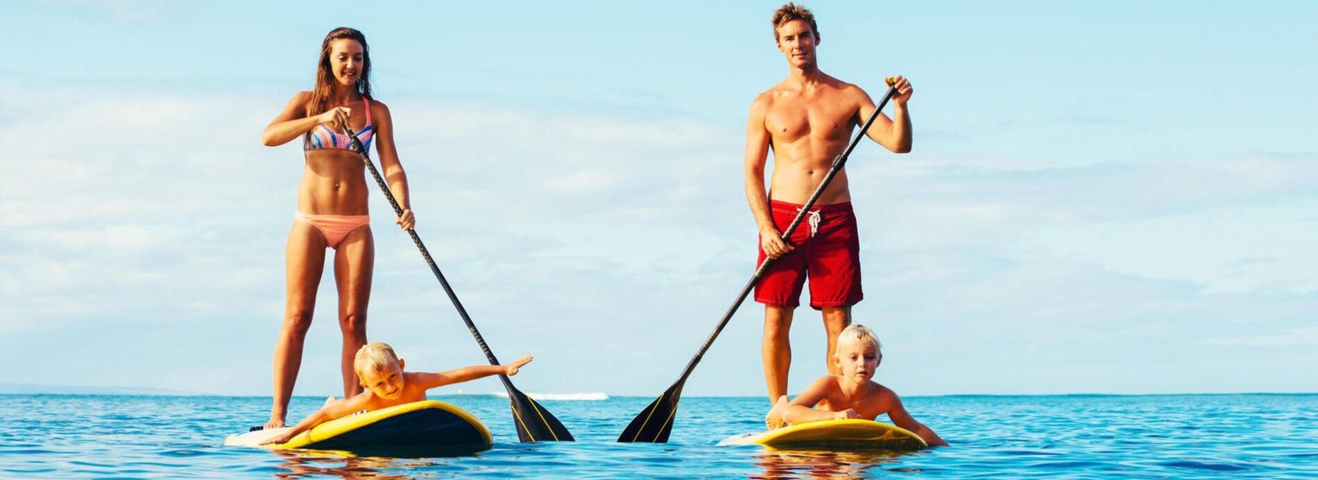 A man standing on a paddleboard holding an oar while a young child rests on a board in smooth blue ocean water.