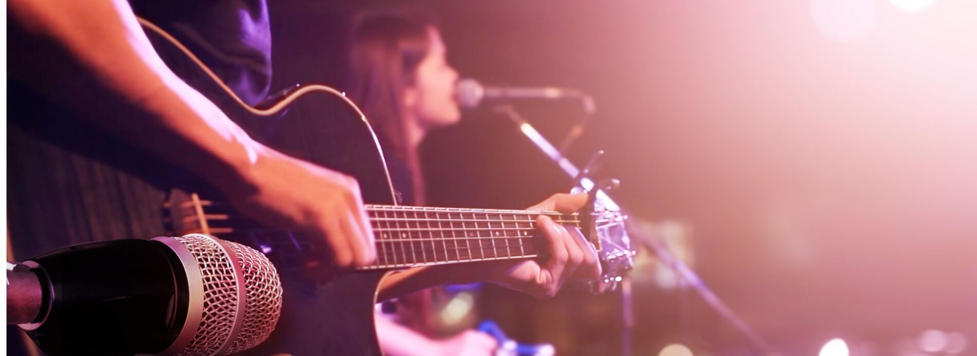 Another perspective of a musician singing into a microphone and strumming a guitar under warm purple stage lighting.
