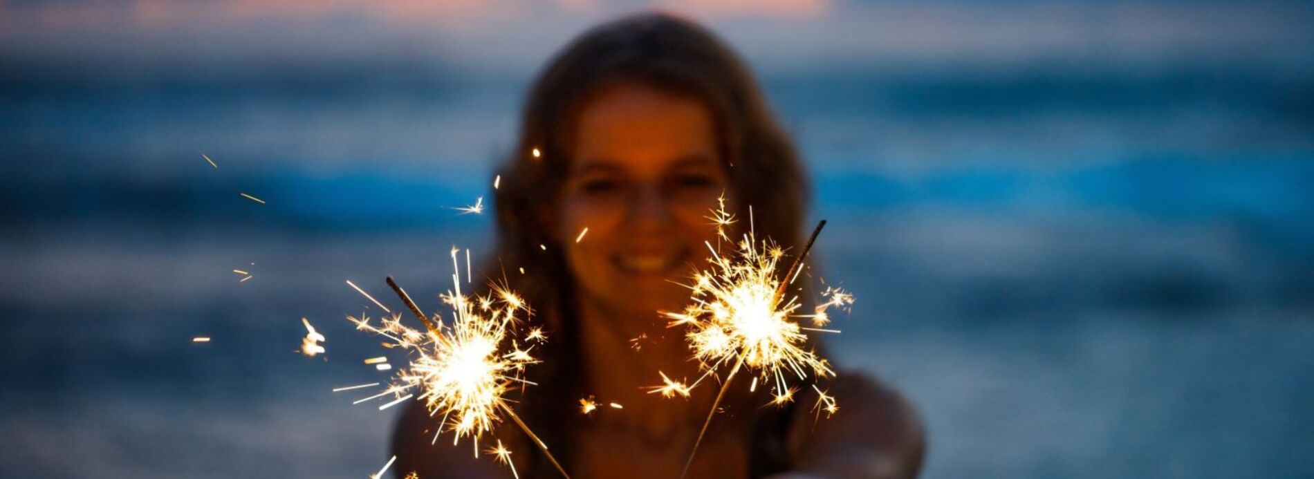 A young woman smiling warmly while holding a lit sparkler outdoors during an evening beach celebration or holiday.