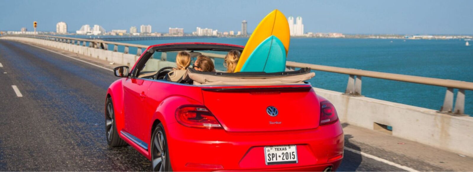 Driving-convertibles-along-the-beach-coast | South Padre Island Vacation Rentals A bright red convertible car with a surfboard packed in the back seat parked near the sand, embodying the ultimate coastal road trip.
