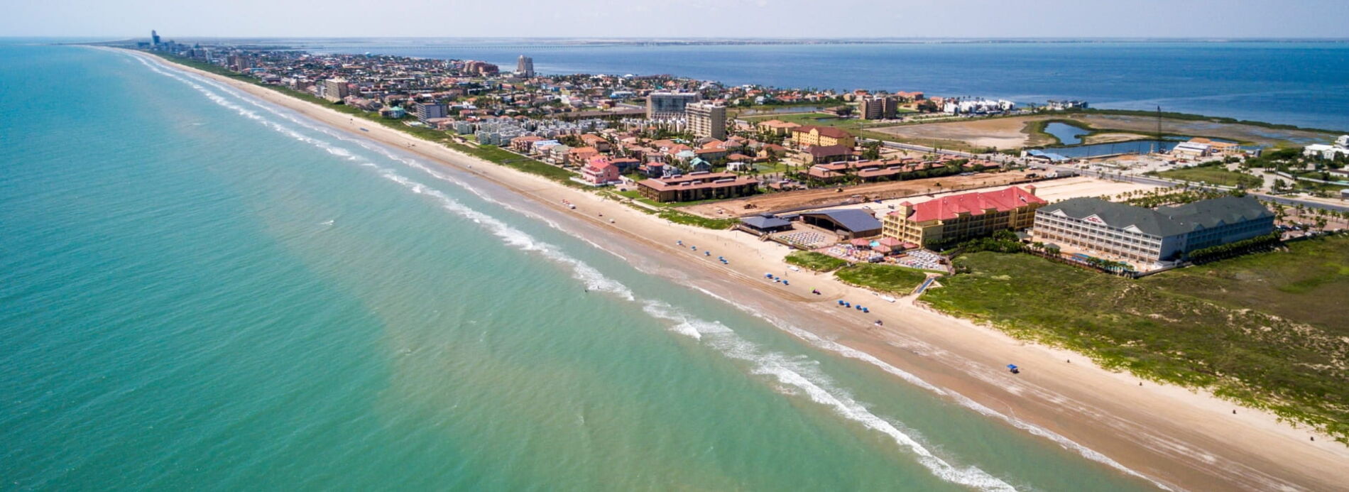 A sweeping aerial view looking down miles of pristine shoreline where light sand meets deep emerald-green ocean surf.