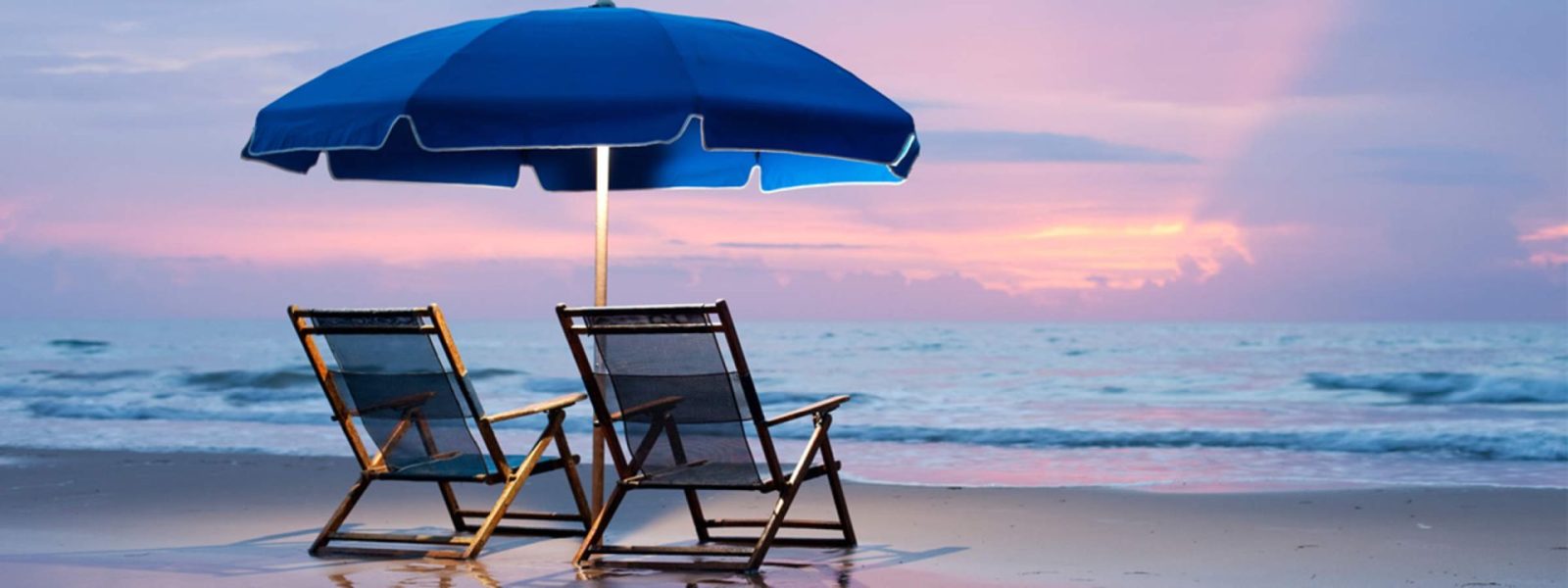 Beach-chairs-and-blue-umbrella-setup | South Padre Island Vacation Rentals A single empty beach lounge chair nestled beneath a royal blue sun umbrella facing the peaceful ocean waves.